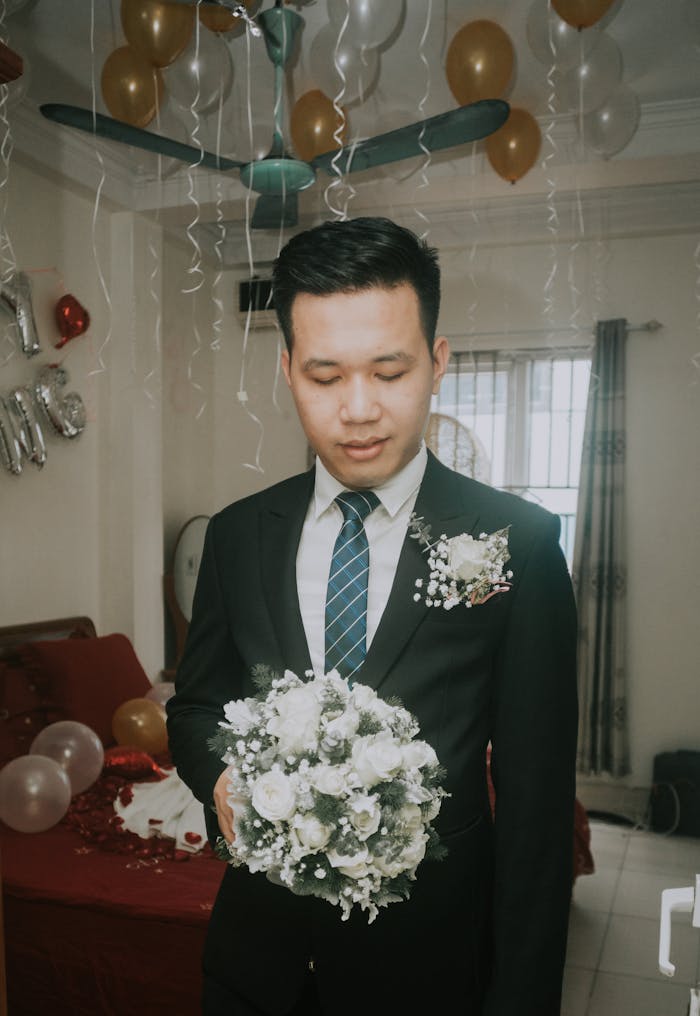 A groom in a black suit holding a white bridal bouquet indoors with balloons.